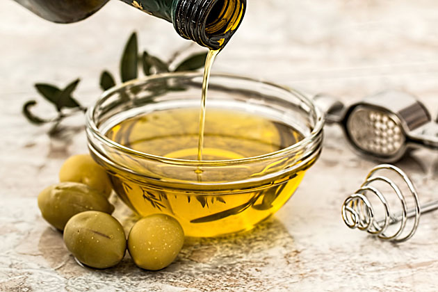 Abbildung 1: Olivenöl wird aus einer Flasche in eine Schale gegossen, daneben liegen grüne Oliven (Archivbild, Pexels, CC0 Public Domain).